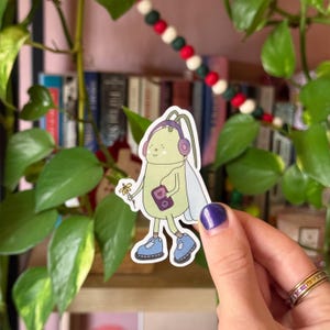 May include: A green and yellow cartoon bug wearing headphones and holding a small purple and pink cassette player. The bug is wearing blue shoes and has a yellow flower in its hand.