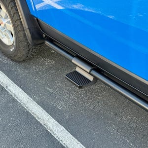 May include: A black side step attached to a bright blue truck. The step is rectangular with a ridged surface for traction. A black bar runs along the side of the vehicle, and a portion of the tire is visible.