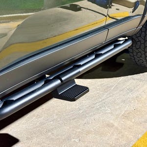 May include: Black side steps attached to a vehicle. The steps are made of a tubular metal with a textured step pad. The vehicle's tire and body panel are visible in the image.