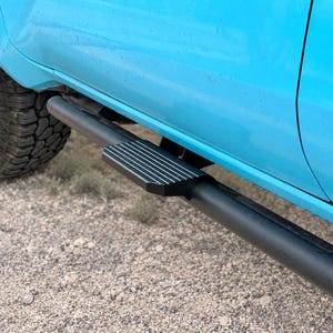 Puede incluir: Un estribo tubular negro mate con una almohadilla texturizada y estriada, montado debajo del panel de la puerta azul brillante de un vehículo. El estribo cuenta con una barra principal cilíndrica y una sección rectangular con ranuras de agarre.