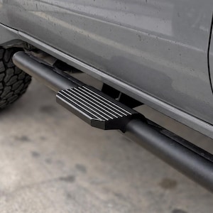 Puede incluir: Un estribo tubular negro mate con una almohadilla texturizada y estriada, montado en el lateral de un vehículo gris. El escalón de metal presenta un diseño de superficie con ranuras horizontales y está fijado al chasis bajo la puerta.