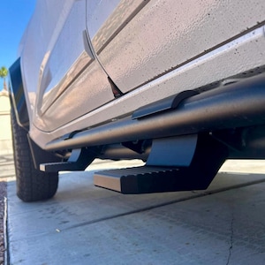 May include: Black side steps attached to a white truck. The steps are rectangular with a textured surface for grip. The running board is a black tube that runs the length of the truck.