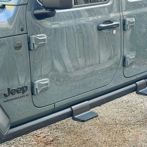 Può includere: Primo piano di una portiera grigia di Jeep Wrangler. La portiera presenta una maniglia nera, cerniere e un gradino. Il logo Jeep Wrangler è visibile. Il veicolo è parcheggiato su una superficie di ghiaia.