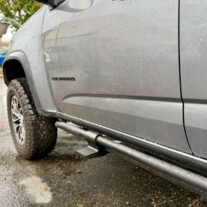 May include: A gray pickup truck with black side steps and a black tire. The word "COLORADO" is visible on the side of the truck. The truck is parked on a wet surface.