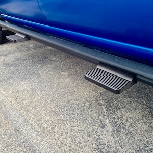 Puede incluir: Estribos laterales negros fijados a un vehículo azul. Los estribos son rectangulares con una superficie estriada para la tracción. El neumático del vehículo es visible en el fondo.