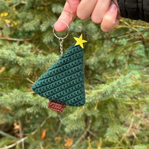 May include: A handmade green crochet Christmas tree keychain with a yellow star on top and a brown trunk. The keychain is attached to a silver ring. The background is a blurred view of green pine needles.