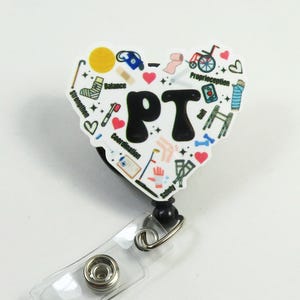 May include: A white heart-shaped retractable badge holder with a black and white design featuring the letters "PT" and various physical therapy related icons. The icons include a wheelchair, crutches, a walker, a cane, a sock, a bandage, and the words "balance", "strength", "coordination", and "proprioception".