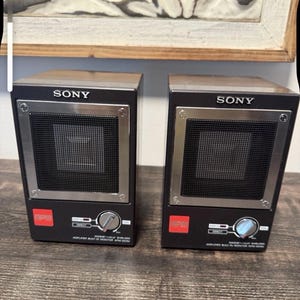 May include: A pair of black Sony APM-007AV magnetically shielded monitor speakers with built-in amplifiers. Each speaker features a square black mesh grille, a silver volume control knob, a red logo badge, and white text labels on the front panel.