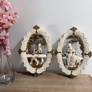 May include: Two oval wall plaques featuring cream-coloured frames with gold-toned accents. Each plaque depicts a three-dimensional scene of a figure sitting on a bench under a tree. One figure plays a stringed instrument, while the other sits in a long gown.