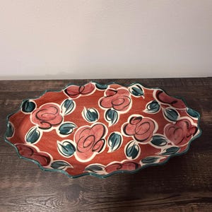 May include: An oval ceramic serving platter with a scalloped edge, featuring a terracotta-colored background decorated with hand-painted stylized pink roses and dark teal leaves outlined in black, creating a bold and expressive floral pattern.