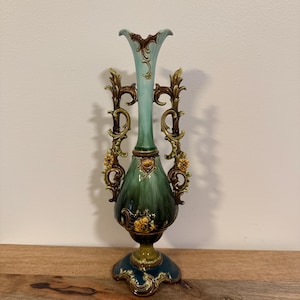 May include: A tall ceramic vase featuring a gradient color scheme transitioning from light blue at the top to dark green and deep blue at the base. The vase has two ornate, scroll-like handles with yellow floral accents and a flared rim.