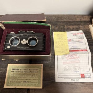 May include: A vintage View-Master Personal Stereo Camera in its original box. The black camera has two lenses and a silver dial. Included is a warranty card and a receipt from a photo shop. The box is open, revealing a maroon interior.