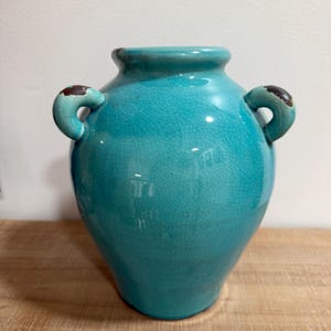May include: A turquoise ceramic vase with two curved handles. The vase has a crackle glaze finish and a slightly distressed look. The handles have a dark brown accent. The vase is sitting on a wooden surface.
