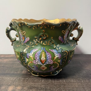 May include: A dark green ceramic vessel with two ornate handles and a scalloped rim. The exterior features intricate gold filigree scrollwork and symmetrical floral motifs in shades of purple, blue, and orange. The interior has a gold finish.