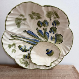 May include: A white ceramic serving platter with a green rim and a leaf-like shape. The platter is decorated with hand-painted blue and green vegetables, including carrots, mushrooms, and asparagus. A smaller bowl is nested inside.