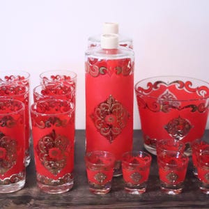 Puede incluir: Un juego de cristalería con un acabado esmerilado en rojo vibrante y patrones de filigrana dorada. La colección incluye una jarra alta con tapón, una cubitera ancha, varios vasos altos para beber y una serie de vasos pequeños para chupitos.