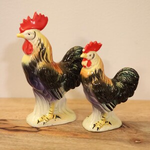 May include: Two ceramic rooster figurines of different sizes, each featuring a bright red comb and wattle, a yellow neck, and a body with black, brown, and purple plumage. Both birds stand on a cream-colored base with yellow legs and black claws.