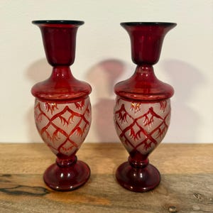 Peut inclure: Une paire de bougeoirs en verre rouge rubis avec un sommet évasé, un col fin et une base bulbeuse. La partie inférieure de chaque bougeoir présente une finition givrée avec un motif en treillis rouge foncé en relief rappelant des feuilles.
