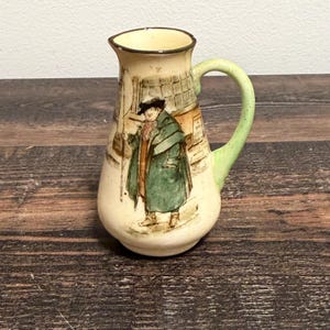 以下が含まれることがあります： ダークブラウンの縁とライトグリーンのハンドルが付いたクリーム色の陶器製ピッチャー。ピッチャーの本体には、長い緑色のコートと黒い帽子を身に着け、建物の前に立っている人物の精巧なイラストが描かれています。