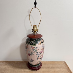 May include: A table lamp with a ceramic base featuring a pale pink background decorated with colorful floral patterns and green foliage. The lamp has a dark red circular lid and base, a brass harp, and a black finial at the very top of the structure.