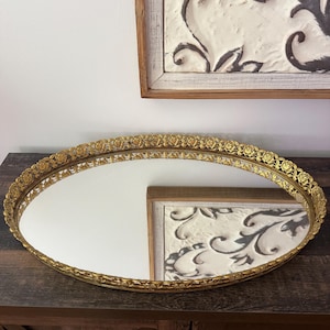 Puede incluir: Una bandeja ovalada con una base de espejo reflectante y un borde decorativo de metal dorado. El borde elevado está elaborado con un patrón repetitivo de pequeñas rosas detalladas y complejos recortes de filigrana a lo largo del contorno.