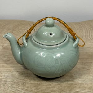 Puede incluir: Una tetera de cerámica en color verde celadón claro con un diseño de pez dorado tallado en relieve en el cuerpo. La tetera tiene una tapa a juego con un pequeño orificio para el vapor y un pico curvo. Un asa de bambú trenzado marrón.