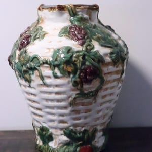Può includere: Un vaso in ceramica con una texture a intreccio di cesto bianco, caratterizzato da foglie di vite verdi in rilievo dipinte a mano e grappoli d'uva viola scuro. Il bordo e la base presentano accenti marroni e la superficie è lucida.