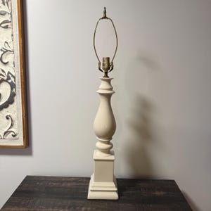 May include: A tall, off-white table lamp base featuring a smooth, curved, baluster-style central column that transitions into a square, tiered pedestal base. The lamp includes a brass-colored metal socket and a wire harp for shade attachment.