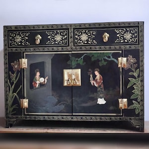 May include: A black rectangular cabinet featuring two top drawers with gold floral accents and two central doors. The doors display painted figures in a garden setting, complemented by gold-toned metal hinges, drawer pulls, and latch hardware.