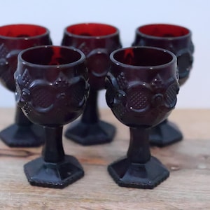 Könnte beinhalten: Ein Set aus fünf tiefroten Glas-Kelchen mit einem erhabenen geometrischen Muster auf der Kuppa, einem kurzen Stiel und einem sechseckigen Fuß. Das Glasgeschirr hat ein dunkles, durchscheinendes Aussehen mit einem strukturierten Oberflächendesign.
