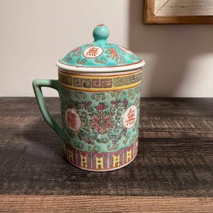 May include: A ceramic mug with a lid, featuring a light green background, a central pink floral motif, and red Chinese characters. The design includes a yellow geometric border near the top and a colorful vertical pattern along the base.