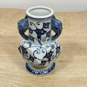 Op de afbeelding: Een witte keramische vaas met handgeschilderde blauwe bloemmotieven en ranken. De vaas heeft een cilindrische hals, een bolvormig lichaam met gefacetteerde zijkanten en twee gebeeldhouwde handvatten in de vorm van dierenkoppen. De voet is versierd met een blauwe zigzagrand.