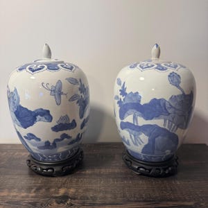 May include: A pair of white ceramic ginger jars with blue hand-painted floral and butterfly motifs. Each jar features a removable lid with a pointed finial and rests on a dark brown carved wooden base, showcasing detailed botanical patterns.