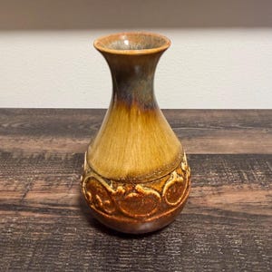 May include: A ceramic vase featuring a flared rim and a tapered neck. The upper portion has a smooth, glossy honey-brown glaze that transitions into a darker, textured base adorned with raised, swirling scrollwork patterns in deep brown tones.