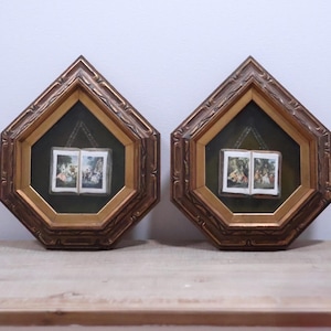 May include: A pair of pentagonal wall frames with ornate gold-toned edges and dark green velvet backing. Each frame contains a small, open book display featuring miniature colour illustrations of figures in outdoor settings, centred behind glass.