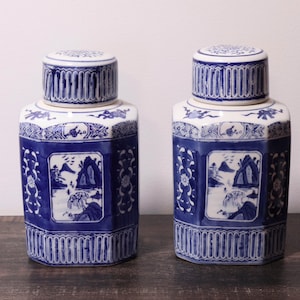 Peut inclure: Une paire de pots à gingembre en céramique hexagonaux présentant une base blanche avec des paysages peints à la main en bleu, des motifs floraux et des bordures géométriques. Chaque pot inclut un couvercle amovible assorti décoré.