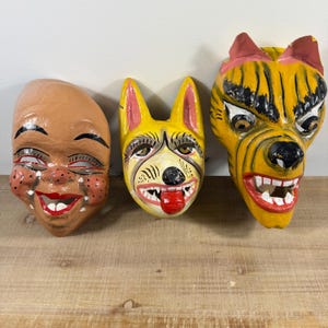 以下が含まれることがあります： 手描きのマスク3点。赤い頬をしたベージュの人の顔、赤い舌を出した黄色い犬の顔、ピンクの耳をした黄色い虎のような生き物の顔。それぞれのマスクには、描かれた目や歯など、詳細な顔の特徴が表現されています。