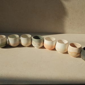 Op de afbeelding: Een rij van tien kleine, ronde keramische bekers in verschillende tinten groen, wit en bruin. De bekers zijn in een rij op een lichtbruin oppervlak gerangschikt.