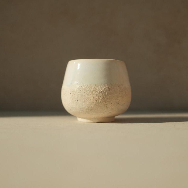 Op de afbeelding: Een witte keramische beker met een textuur, gespikkeld oppervlak. De beker heeft een ronde bodem en een brede, licht uitlopende opening.