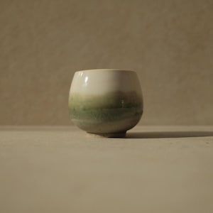 Op de afbeelding: Een witte keramische beker met een groene en bruine glazuur. De beker heeft een afgeronde vorm en een brede basis.