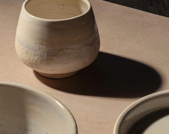 May include: Three ceramic bowls in various sizes, all in a creamy white color with a speckled texture. The bowls are arranged on a light brown surface.