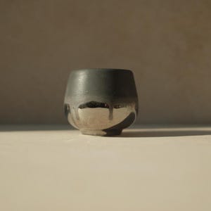 Op de afbeelding: Een kleine, ronde, grijze keramische beker met een zilveren glazuur op de onderste helft. Het glazuur heeft een druppel-effect, waardoor een unieke en artistieke uitstraling ontstaat.