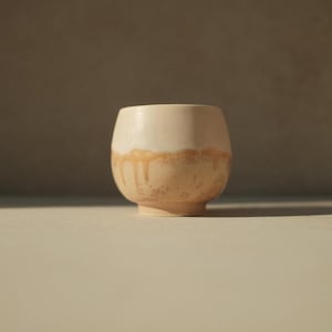 Op de afbeelding: Een keramische beker met een witte glazuur en een bruine druppelglazuur. De beker heeft een afgeronde vorm en een brede basis.