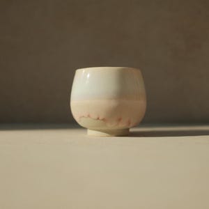 Op de afbeelding: Een keramische beker met een lichtblauwe glazuur en een roze rand. De beker heeft een ronde vorm en een klein voetje.