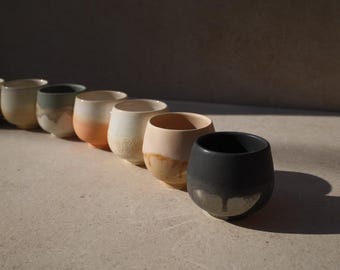 May include: A row of ten ceramic cups in various shades of white, gray, and black. The cups are arranged in a line from left to right, with the largest cup on the right and the smallest cup on the left. The cups are all made of a smooth, glazed ceramic.