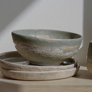 Op de afbeelding: Een lichtgroene keramische kom met een getextureerde rand en een witte glazuur. De kom staat op twee beige keramische borden.