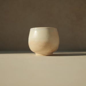 Op de afbeelding: Een witte keramische beker met een getextureerd, licht iriserend glazuur. De beker heeft een afgeronde vorm en een brede basis.