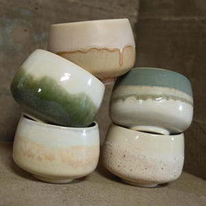 May include: A stack of four ceramic bowls with different glaze patterns. The top bowl is a light beige with a brown drip glaze. The second bowl is a white bowl with a green and white glaze. The third bowl is a white bowl with a blue and white glaze. The bottom bowl is a white bowl with a speckled brown glaze.