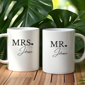 May include: Two white ceramic mugs with black text. One mug reads "MRS. 2026 Johnson" and the other reads "MR. 2026 Johnson". The mugs have a curved handle and are set against a background of green foliage.