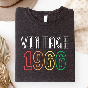 Könnte beinhalten: Dunkelgraues T-Shirt mit dem Text "VINTAGE 1966" in Weiß, Rot, Orange, Gelb und Grün. Das gefaltete Shirt liegt auf einer weißen Oberfläche, mit einem Hut und dekorativem Stoff im Hintergrund.